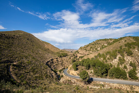 View of Puerto de la Cadena with winding road between arid mountains with low scrub and pine trees and intense blue sky