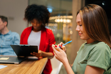 Close-up side view of pretty smiling young woman taking notes on meeting sitting at table during multicultural professional businesspeople working together on research plan in boardroom, close up.