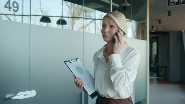 Smiling mature woman wearing formal shirt negotiating with business partners contract terms by mobile phone, walking holding clipboard with presentation deck in corporate business office, slow motion