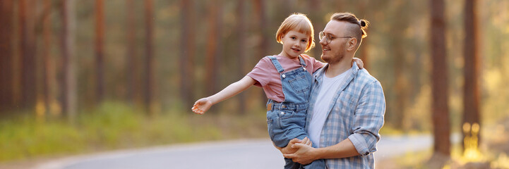 Fototapeta premium Father holds little daughter in his arms and they laugh joyously while strolling along a forest road, surrounded by majestic pine trees. Banner for website header design with copy space.