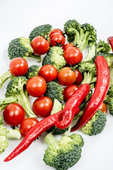 Seen from above, a sizeable amount of red hot chili peppers, ripe cherry tomatoes and raw broccoli stalks on a white surface