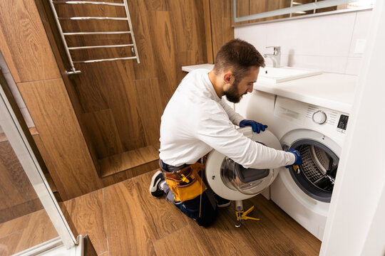 Plumber in overalls with tools is repairing a washing machine in the house.