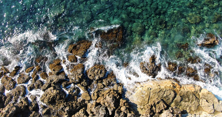 Aerial top view over the island's rocky shore and clear sea with varying shades of blue and light foamy waves in warm summer weather.