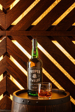 Bottle of Irish whiskey Proper No. Twelve and glass of whiskey on wooden barrel against glowing wall