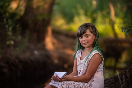 Portrait of girl with green hair sits on the river bank, holding white origami paper boat in hands