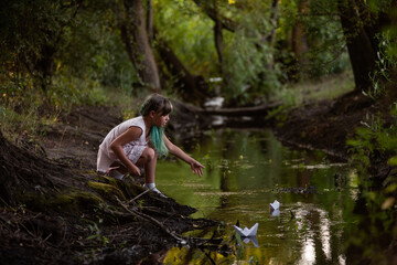 Teenage girl with green hair in dress, stands on the river bank, launches white paper origami boat
