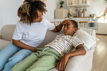 Worried caring African American mom touch forehead of sad child sitting on couch feeling unwell malaise nausea weakness. Attentive black woman mother check temperature of sick unhealthy schoolboy son - Powered by Adobe