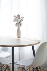 A bouquet of cotton twigs in a vase on a round table with an Easter decor of eggs and an Easter bunny in the light from the window in a minimalist interior.