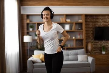 Fototapeta na wymiar Active multiracial woman with dark hair enjoying music in wireless headphones while training at living room. Fitness young lady in sport clothes using modern gadget for workout.