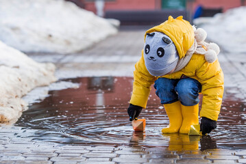 A small child in yellow rubber boots and a jacket runs through puddles, has fun, plays and launches paper boats. Spring break photo. It's springtime.