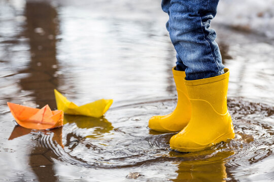 It's springtime. A small child in yellow rubber boots jumps through puddles, plays and launches paper boats on the water.. Photo of spring and autumn holidays. The concept of spring.