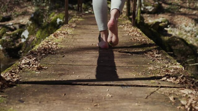 Close-up view of barefoot woman from behind is walking on sunny old wooden bridge. Slow motion.