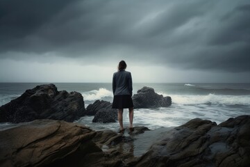 Rear view of businesswoman standing on rock in the ocean facing oncoming waves, created with Generative AI
