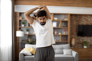 Fototapeta na wymiar Bearded arabian man in sport outfit warming up with arms stretching at bright living room. Flexible exercises for body care and health condition. Domestic training during free time.