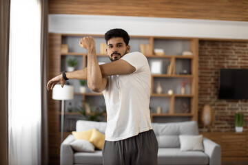 Handsome focused indian man stretching arms for bodybuilder fitness and muscle warm up at cozy living room. Wellness, sport activity and people concept.