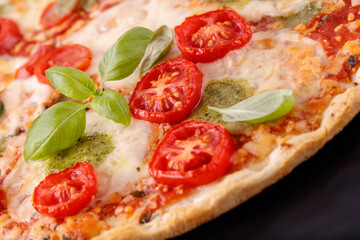 Baked vegetarian crusty pizza with mozzarella, cherry tomatoes, pesto sauce and basil leaves closeup on black bake foil in oven.