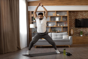 Bearded arabian man dressed in sport clothes standing on yoga mat with hands clenched above head during workout at home. Yoga practice for harmony, balance, soul and body care.