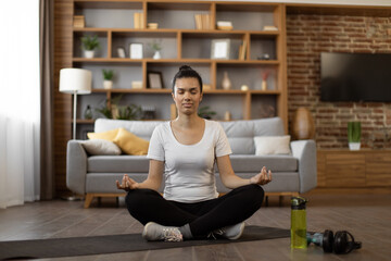 Peaceful interracial woman meditating in lotus pose with closed eyes while sitting on yoga mat....