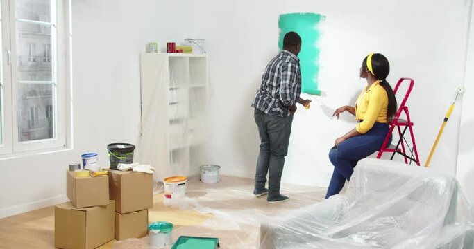 Process of renovation of apartment. Beautiful African-American man painting walls with green paint using roller with caring African woman helping and giving advice. Redesign concept.