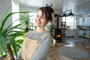 A happy woman in a green house with a potted plant in her hands smiles, takes care of a flower. The interior of a cozy eco-friendly house, a fireplace stove, a hobby for growing and breeding homeplant