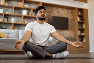 Handsome bearded arabian man in activewear keeping eyes closed and meditating on yoga mat at cozy living room. Concept of chakra, dzen and body wellness.