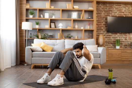 Active indian man in sport clothes lying on yoga mat and training abs with body crunches at home. Fitness workout of strong young guy. Healthy lifestyles concept.