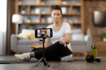 Selective focus of modern gadget on tripod recording active african american woman in sportswear talking at camera while resting on floor indoors. Personal coach guiding fitness training online.