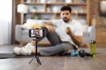 Fototapeta na wymiar Confident sports coach pointing finger at camera and motivating followers to regular physical exercise and active lifestyle. Selective focus of smartphone on tripod filming video content for sharing.