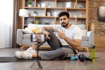 Young indian fitness expert sitting in front of phone camera on yoga mat near sports equipment for home workout. Active male blogger in gym wear recording home exercise for his online blog followers.