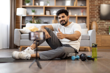 Young indian fitness expert sitting in front of phone on yoga mat near sports equipment for home workout showing thumb up. Active male blogger in gym wear recording home exercise for his online blog