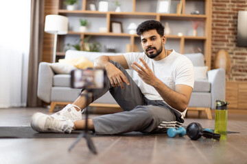 Young indian fitness expert sitting in front of phone camera on yoga mat near sports equipment for home workout. Active male blogger in gym wear recording home exercise for his online blog followers.