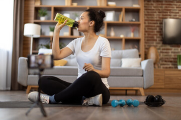 Focused african american female in gym clothes drinking water from sports bottle while sitting cross-legged in front of active cell phone on tripod. Fit sports vlogger showing workout routine at home.