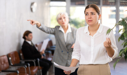 Upset crying young female office worker standing with papers in hands, being scolded by angry woman...