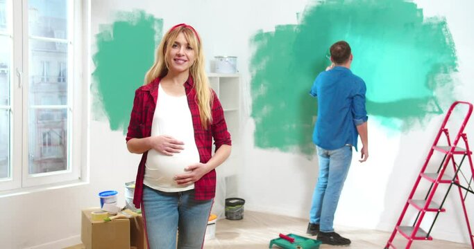 Portrait of happy pretty young Caucasian pregnant woman looking at camera and smiling while handsome loving hard-working husband painting walls using roller on background. Renovation concept.