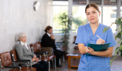 Portrait of smiling young female doctor in blue uniform meeting patient in medical office, filling out medical form at clipboard - obrazy, fototapety, plakaty