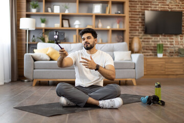 Active indian athlete in gym clothes holding tripod with smartphone and recording video content for sports blog. Attractive bearded male making online tutorial for followers via phone camera at home.