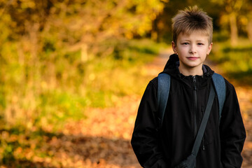 european fair-haired boy in black jacket with backpack on background of orange trees, looking...