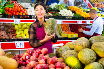 Portrait of smiling confident asian saleswoman standing near counter with fruits at farmers market, offering ripe sweet melon