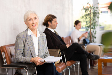 Smiling experienced female entrepreneur sitting in lobby of office with papers in hands, waiting...