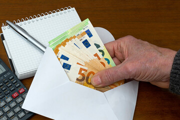 Man's hand holding european banknotes from an envelope. On the table are a calculator and a notepad...