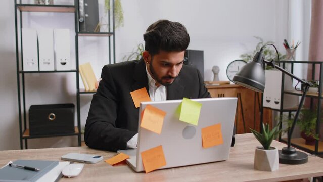 Tired exhausted indian businessman working on laptop at office with many sticker task inscriptions. Freelancer man busy occupation feels sad boredom overworked multitasking. Panic attack, deadline