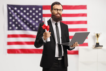 Bearded american man in a suit holding a diploma and a laptop in front of the USA flag