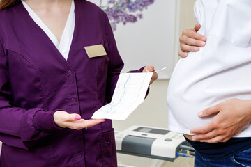 The obstetrician-gynecologist holds the result of the patient's cardiotocography analysis in his hands. A pregnant woman at an appointment with a gynecologist.
