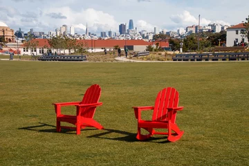 Fotobehang Two red chairs are sitting on green grass at The Presidio's Tunnel Top Park. In the background is the San Francisco cityscape. There is a blue sky with white clouds. © Timothy