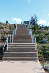 Congrete stairs with metal hand rails. View is looking up to a blue sky. There two platforms. Bushes and plants are each side of the stairs.