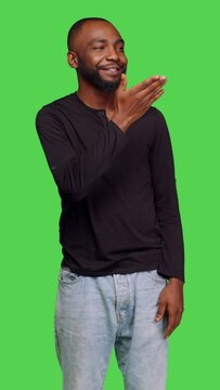 Vertical video: Front view of young person in jeans giving air kisses to people in studio, posing with kissy face on green screen backdrop. Casual male model doing romantic gesture and feeling flirty