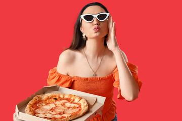 Beautiful woman in sunglasses with box of tasty pizza blowing kiss on red background
