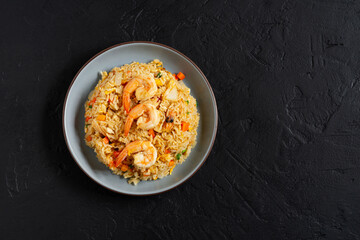 Fried rice with vegetables and sea prawns, carrots, eggs, and tomatoes. Thai Chinese food is arranged on a black plate on a black background kitchen table, top view.
