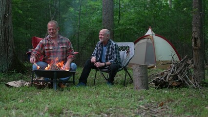 Two gay men building a campfire in a forest talking and laughing with tent in background.