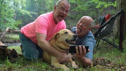 Playful Blonde Labrador Retriever dog sharing love with funny man holding phone taking a selfie in a forest with another man in front of campfire.